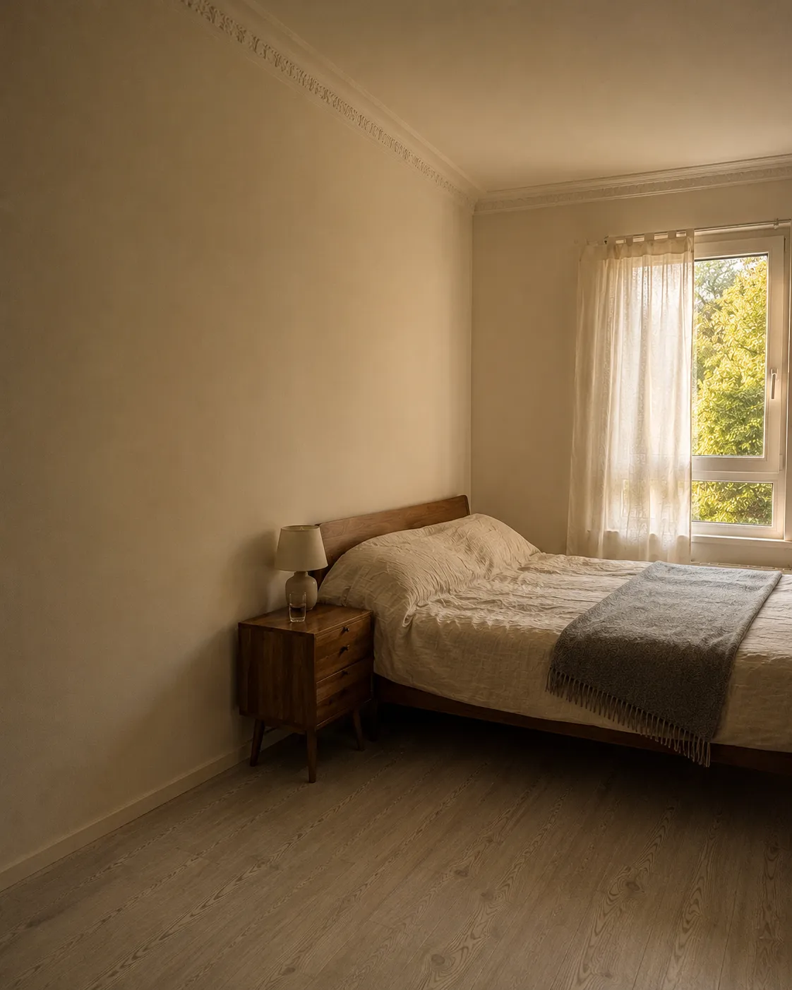 Ein kleines Schlafzimmer mit einem schlichten Holzbett und einem hohen Fenster, im warmen Nachmittagslicht
